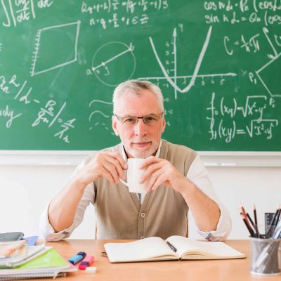 Mathe-Physik Lehrer mit Erfahrung Lehrer mit Kaffeetasse sitzt vor einer Schultafel voller Formeln